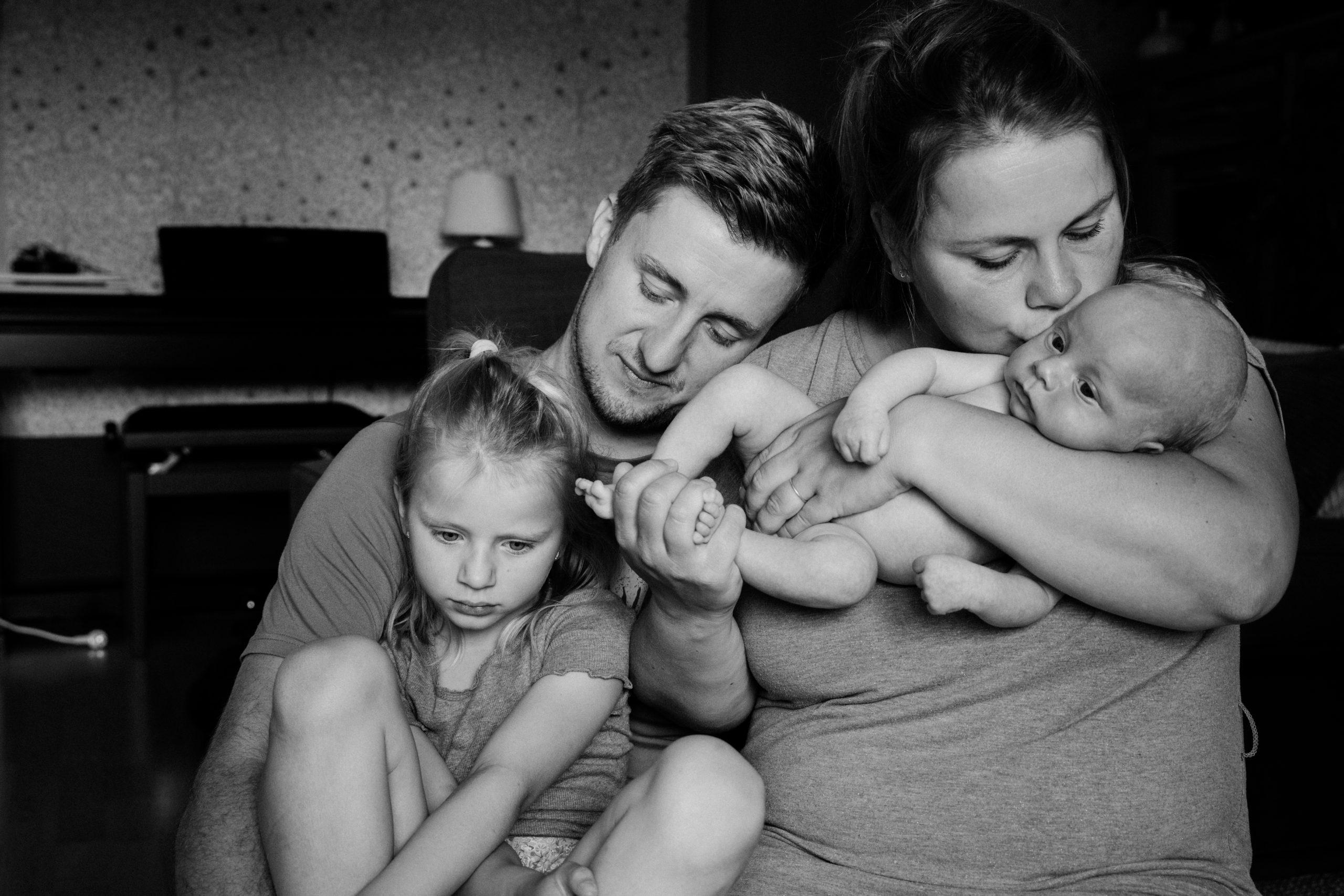 Schwarz-Weiß-Foto: Mutter küsst das Neugeborene, Vater und große Tochter sind dicht dabei