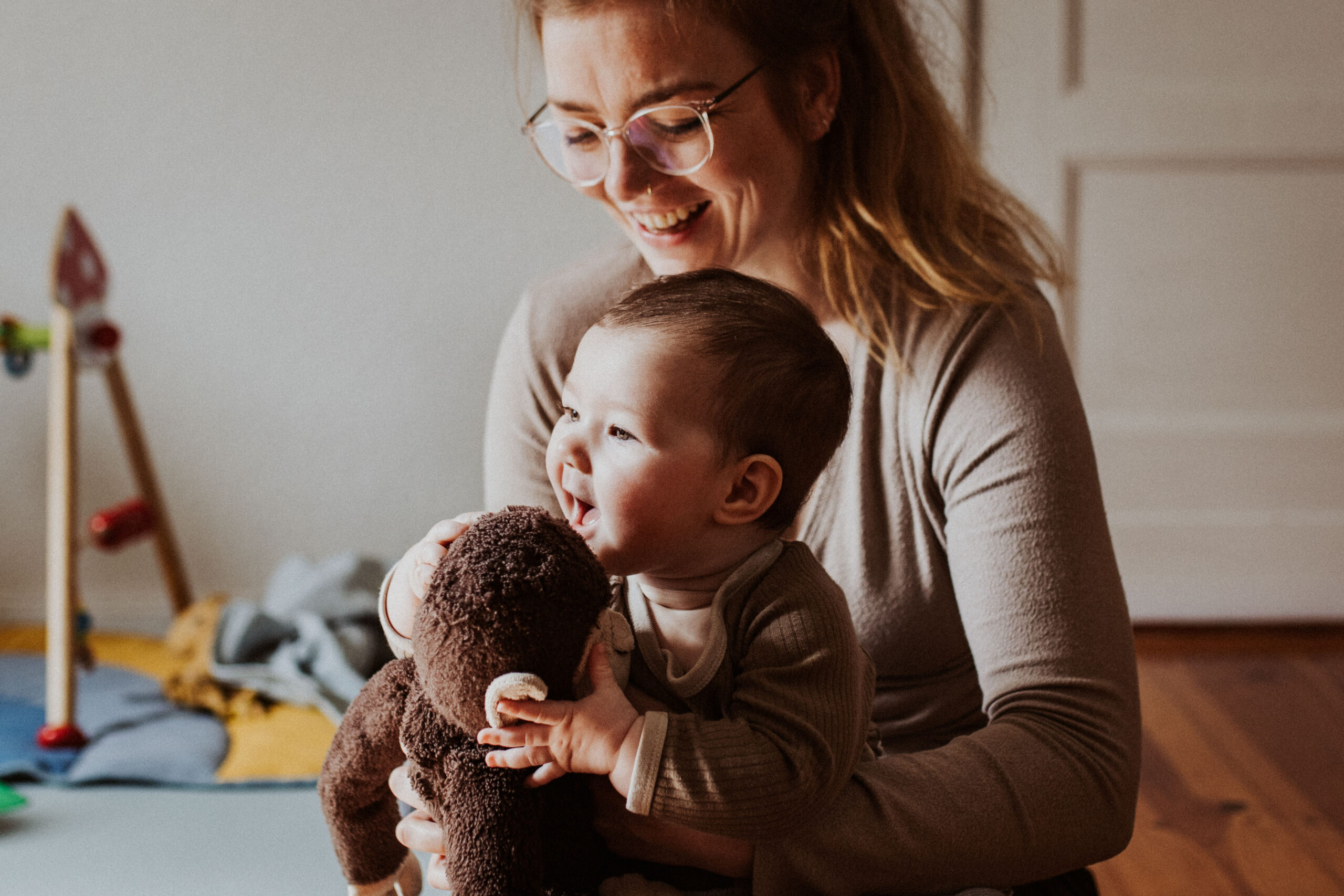 Mutter lacht mit ihrem Baby über ein Kuscheltier auf dem Wohnzimmerboden
